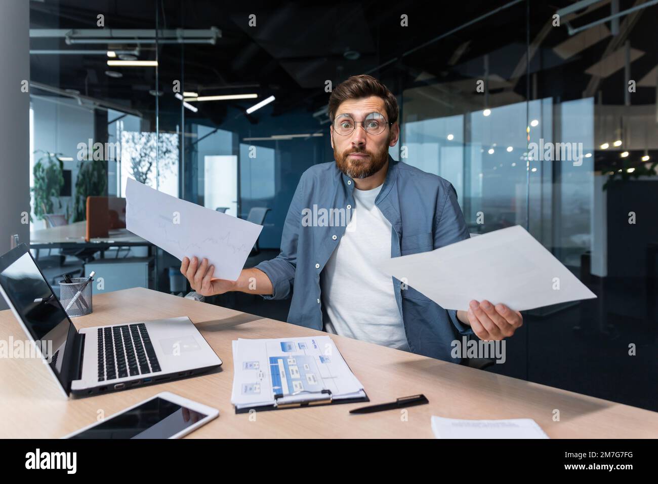 Hombre de negocios insatisfecho y enojado dentro de la oficina que muestra informes y factura documentos a la cámara, jefe en camisa mirando a la cámara trabajando con el trabajo de papel de la computadora portátil. Foto de stock