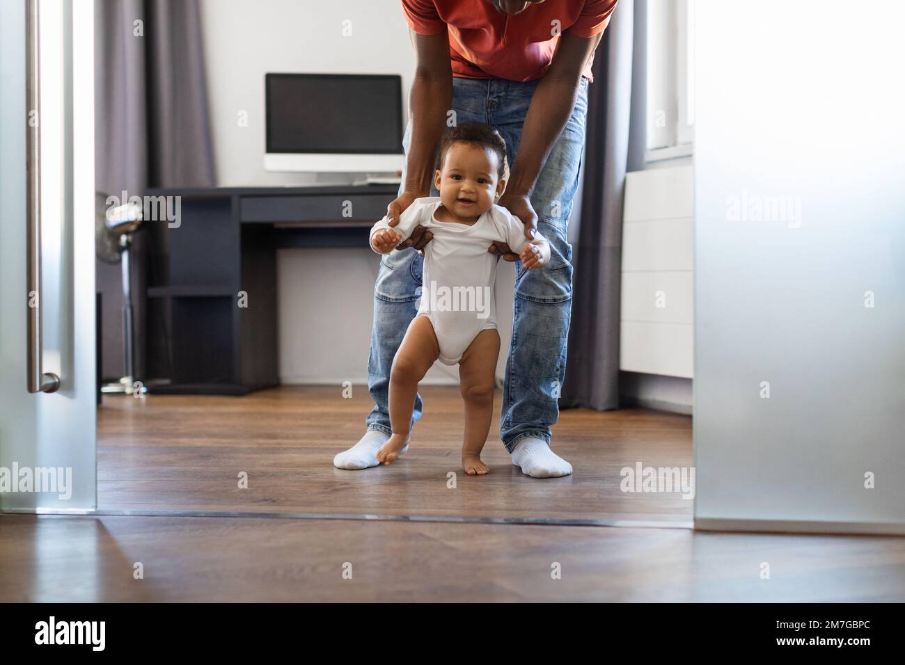 Adorable bebé afroamericano caminando en la sala de estar con la ayuda del padre Foto de stock