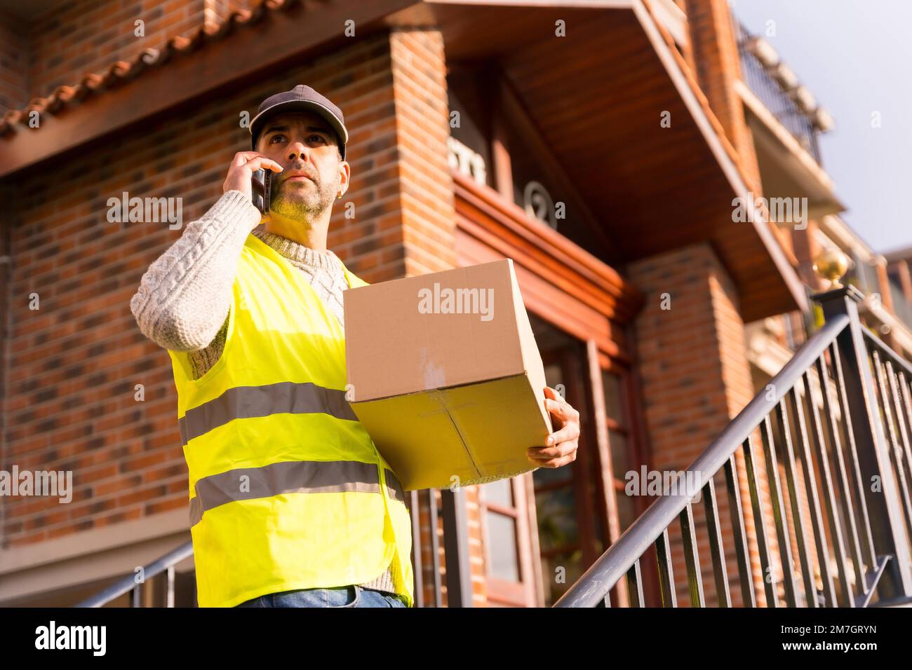 Hombre de entrega de paquetes de una tienda online, con una caja en mano buscando al cliente que llama por teléfono Foto de stock