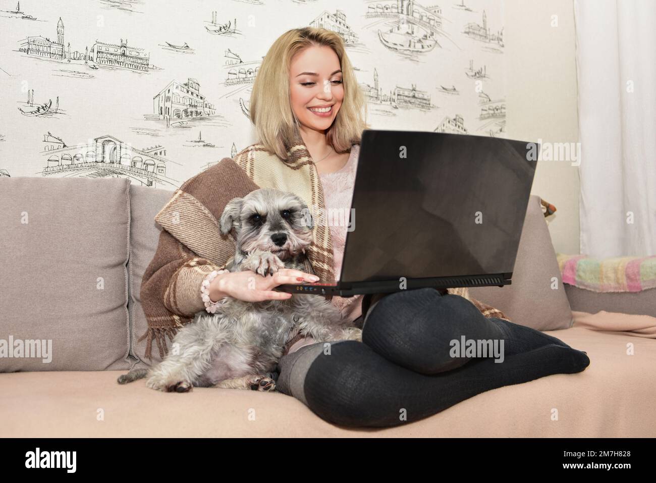 Hermosa mujer rubia sonríe y abraza a un perro lindo en el sofá mientras ve películas en una computadora portátil. Foto de stock