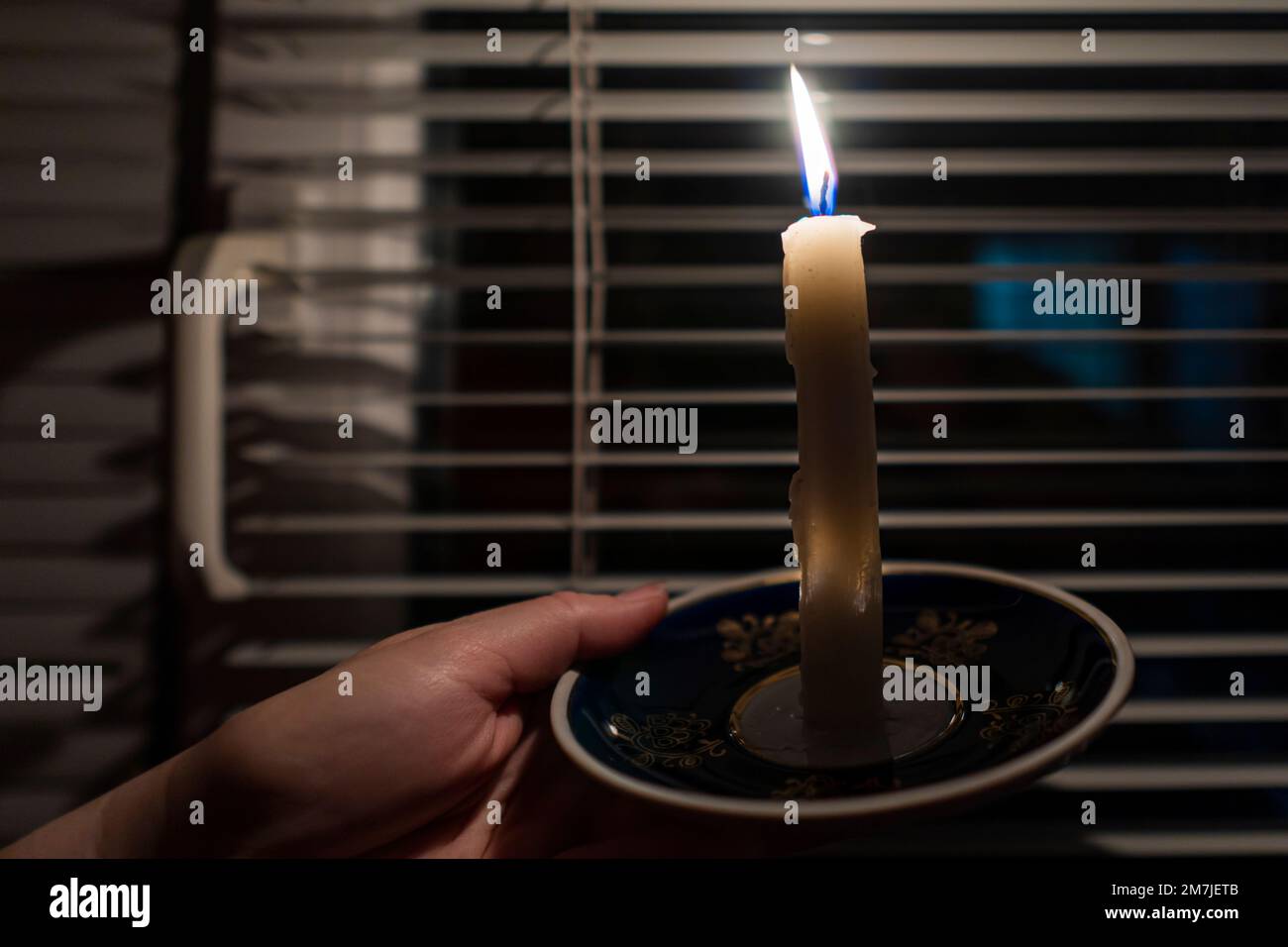 Vela ardiente en un platillo en las manos contra el fondo de una ventana con persianas (primer plano). Concepto de corte de energía. Interrupción. Crisis energética Foto de stock