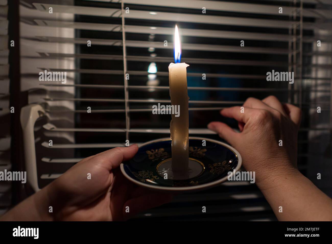 Vela ardiente en un platillo en las manos contra el fondo de una ventana con persianas (primer plano). Concepto de corte de energía. Interrupción. Crisis energética Foto de stock