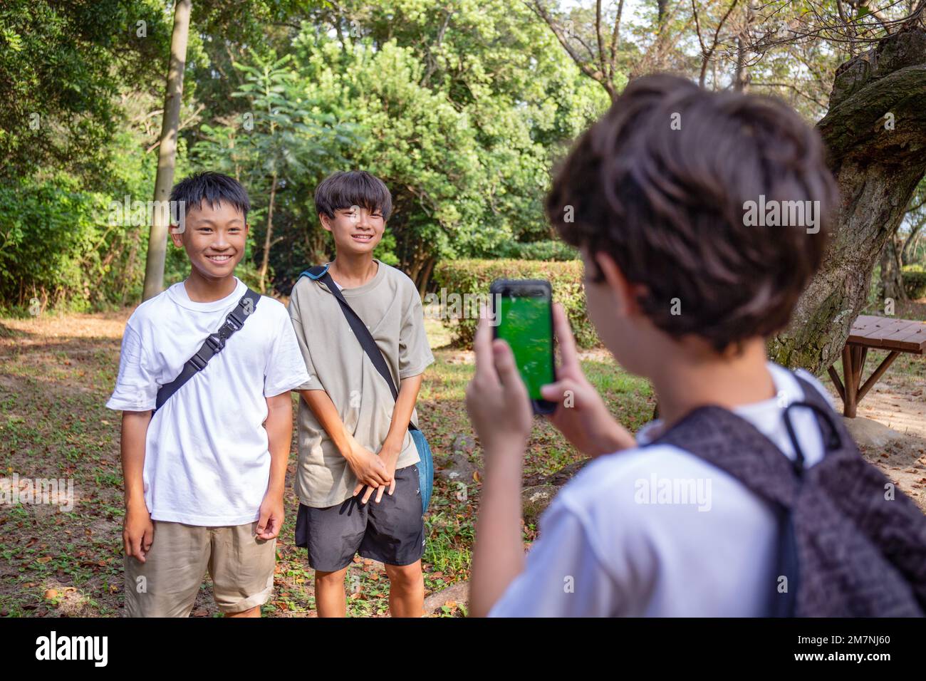 Un niño con un teléfono móvil tomando una foto de dos niños de 13 años uno al lado del otro, al aire libre en un parque en verano. Foto de stock