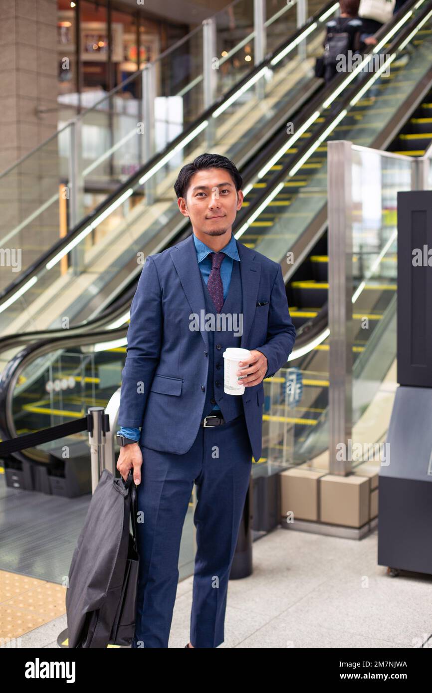 Un joven hombre de negocios con un traje azul en movimiento en un área del centro de la ciudad, llevando un maletín y una taza de café. Foto de stock