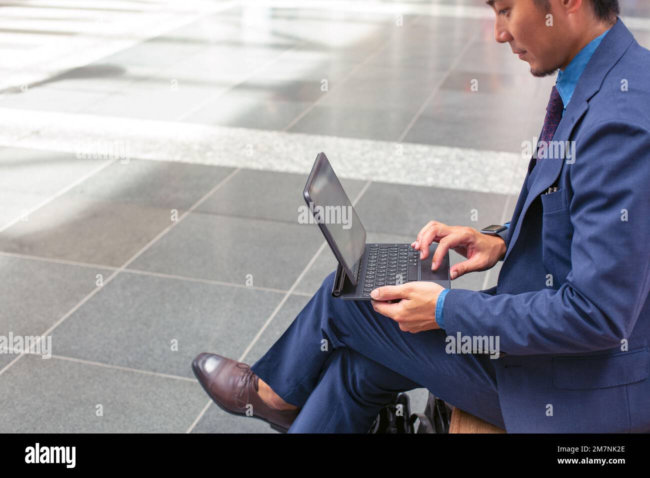 Un joven hombre de negocios con un traje azul en movimiento en un área del centro de la ciudad, sentado en un banco con una tableta digital con una pantalla. Foto de stock
