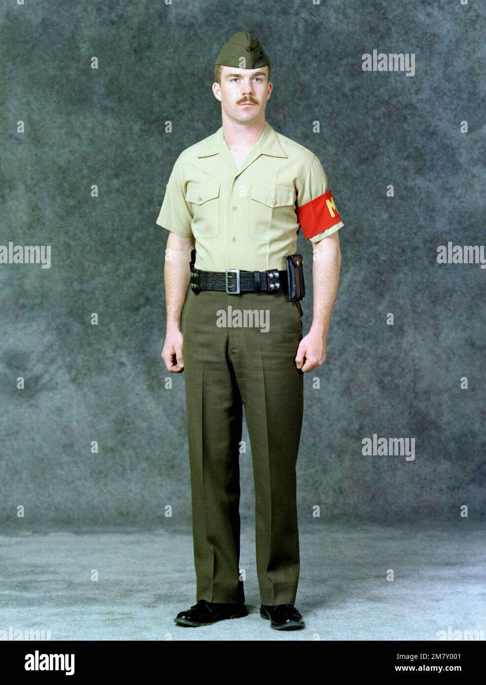 Un Marine SGT. Está vestido con el uniforme de verano de la policía militar  (MP). Lleva pantalones verdes, una camisa caqui, una banda MP para el brazo  y un cinturón de pistola.