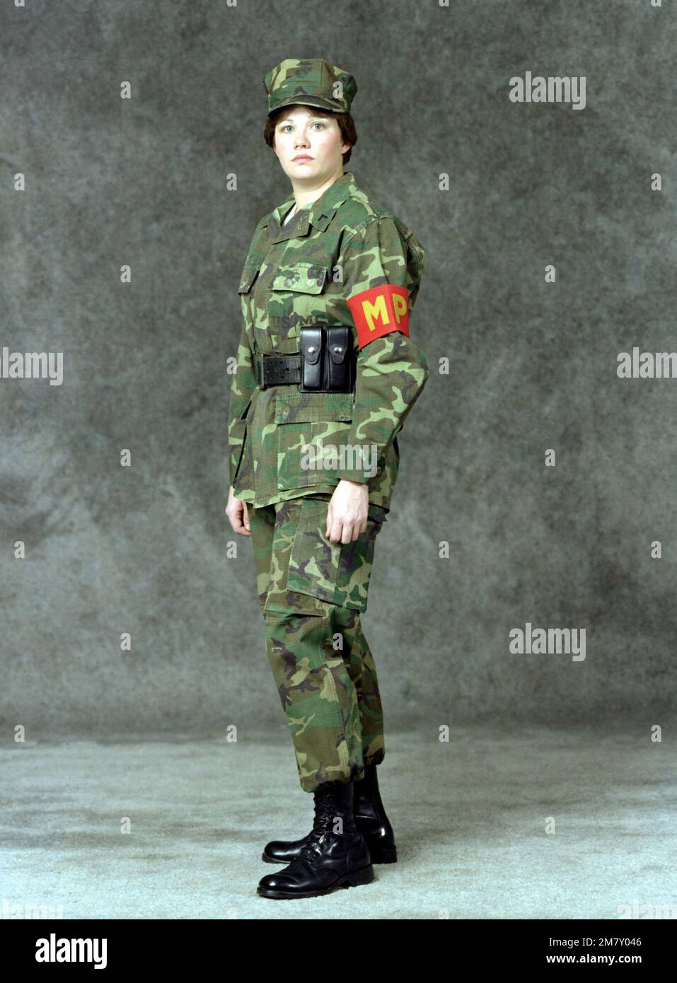 Una mujer de primera clase privada de la Marina está vestida con un  uniforme de trabajo de la policía militar (MP) (peto camuflado). Ella lleva  una banda de brazo MP y un