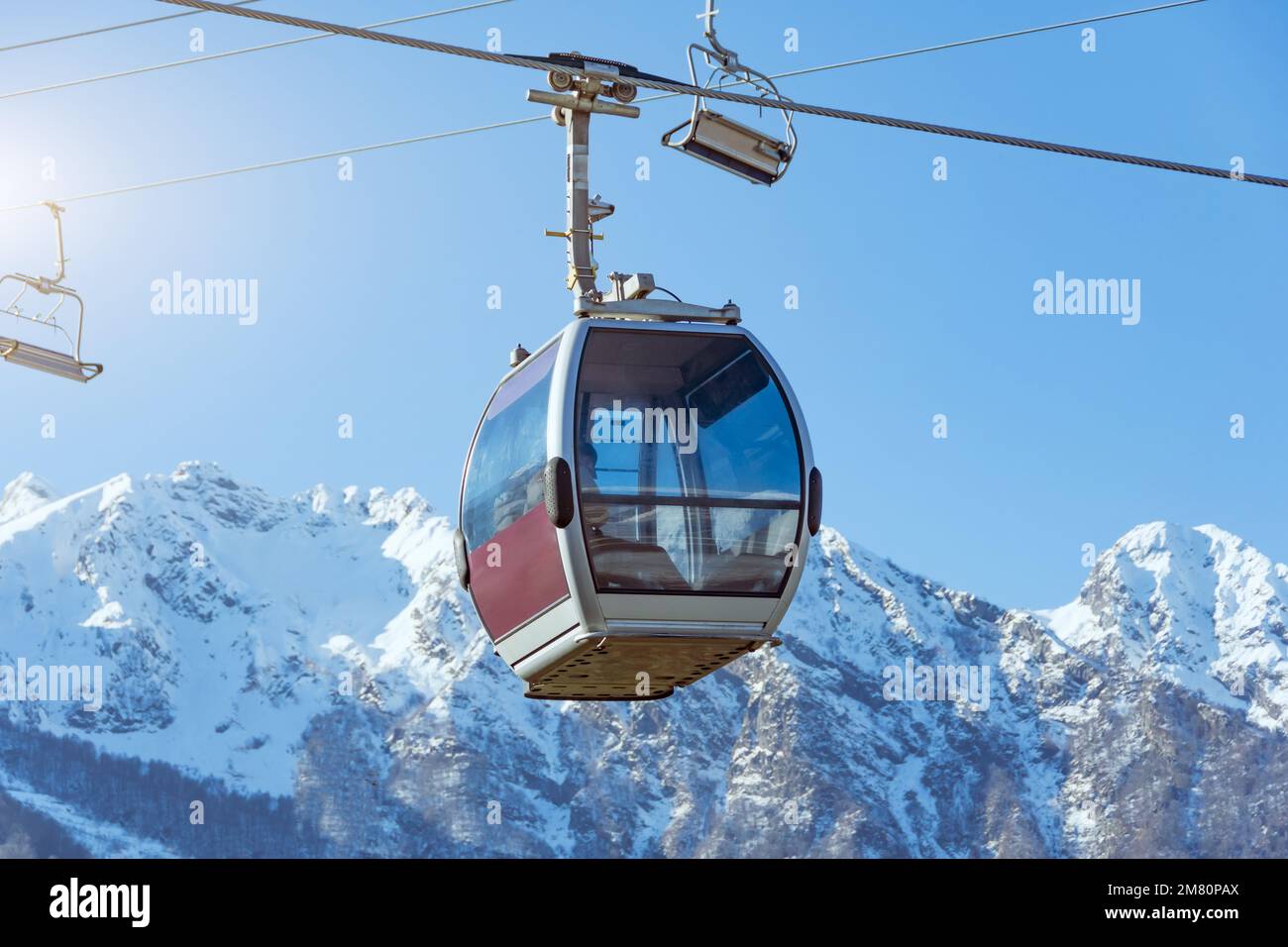Teleférico en las pistas de una estación de esquí con casas y un pico nevado en la distancia, soleado día de invierno Foto de stock