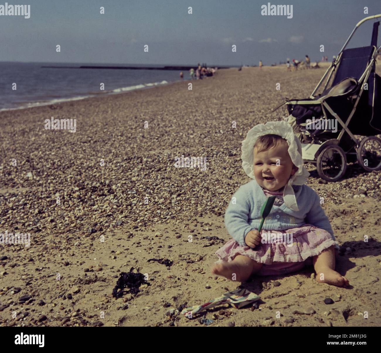 Una niña de un año juega con una pala en Clacton Beach en Essex en 1960. La niña lleva un gorro de algodón blanco para protegerla del sol. La foto está tomada de la diapositiva original. Foto de stock