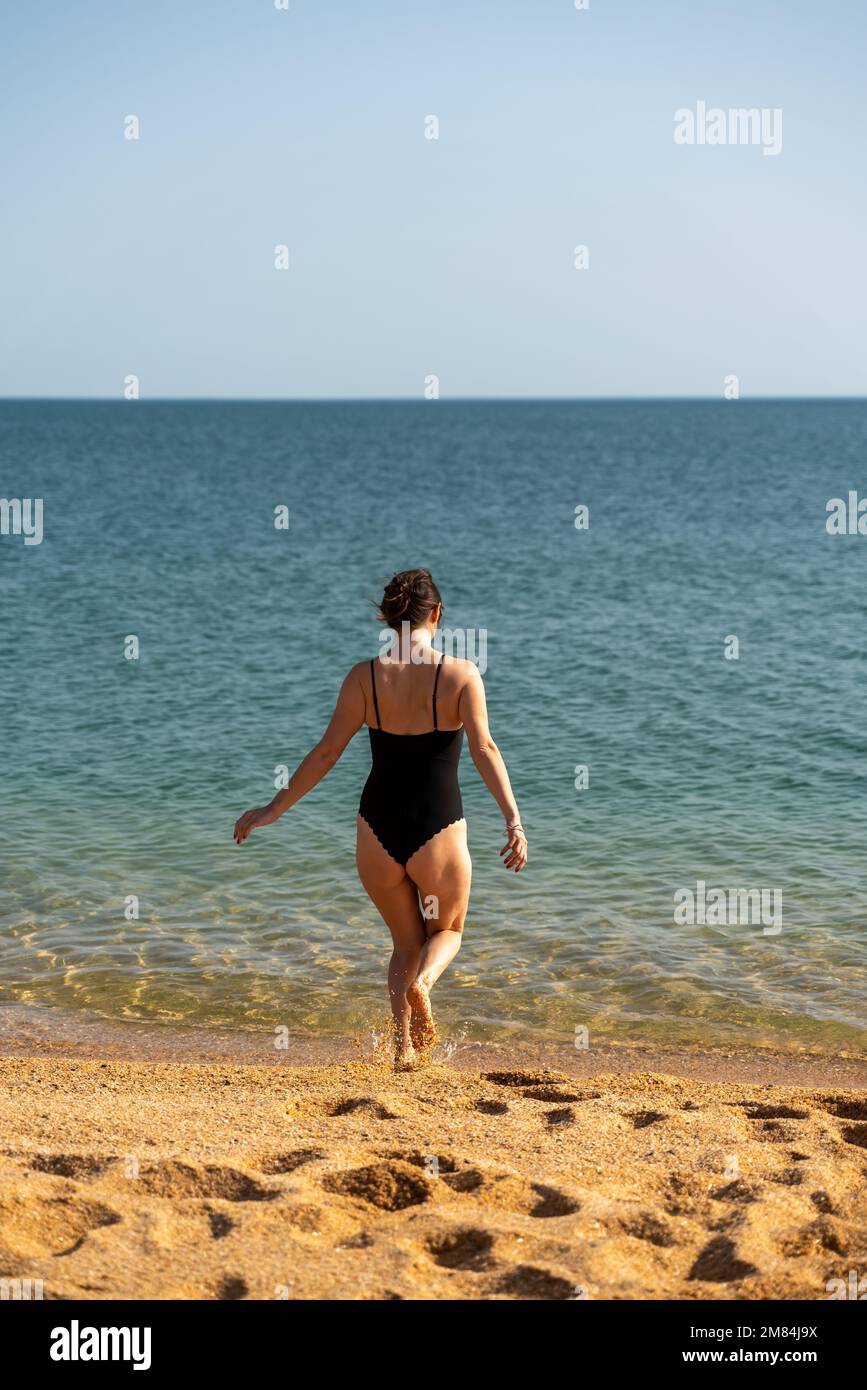 Mujer traje de baño mar arena. Una chica pleno crecimiento se para con su espalda y entra en el en un traje de baño negro. Solo la playa