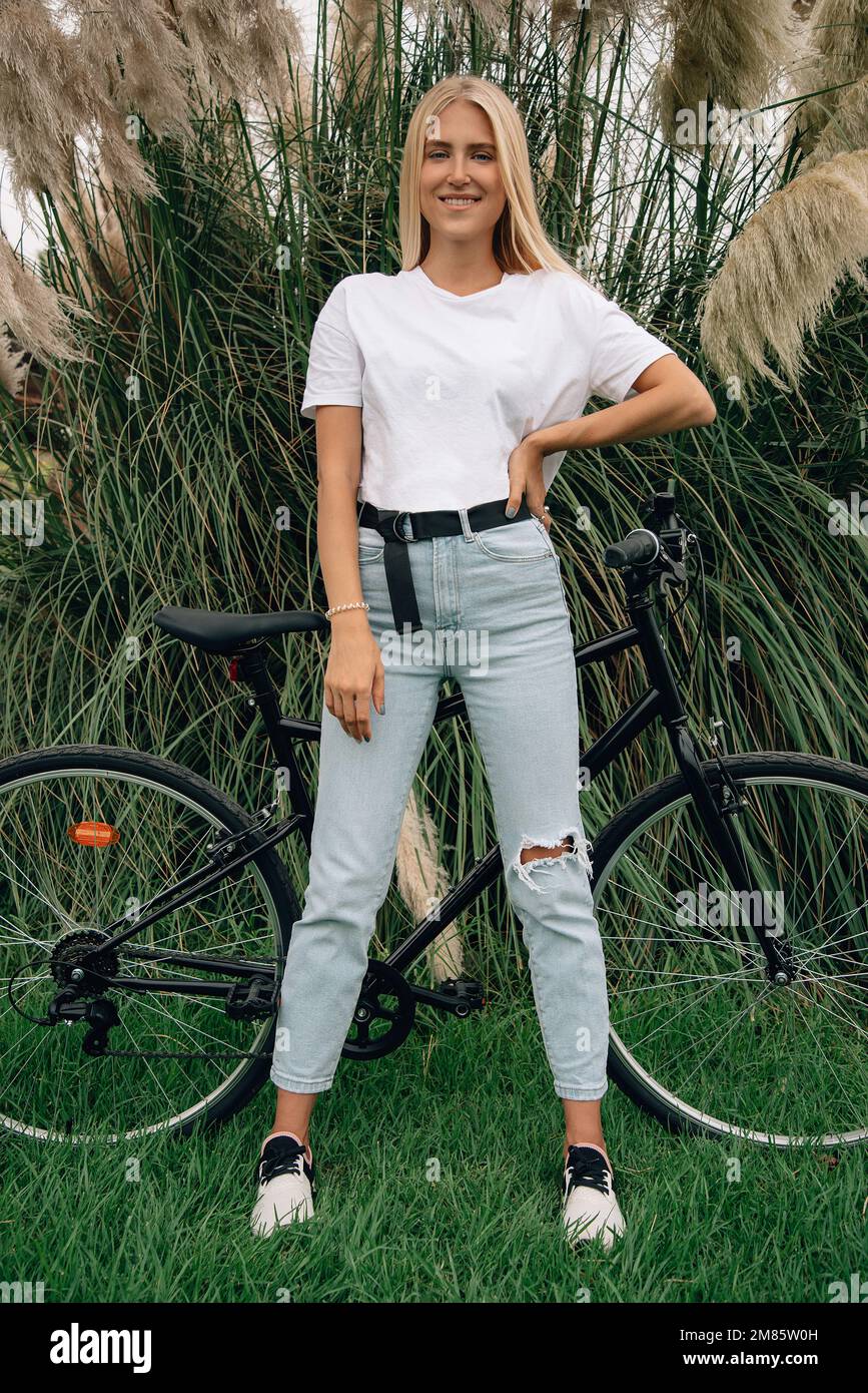 Joven hermosa chica está posando en una camiseta blanca al lado de su bicicleta. Maqueta vertical. Foto de stock