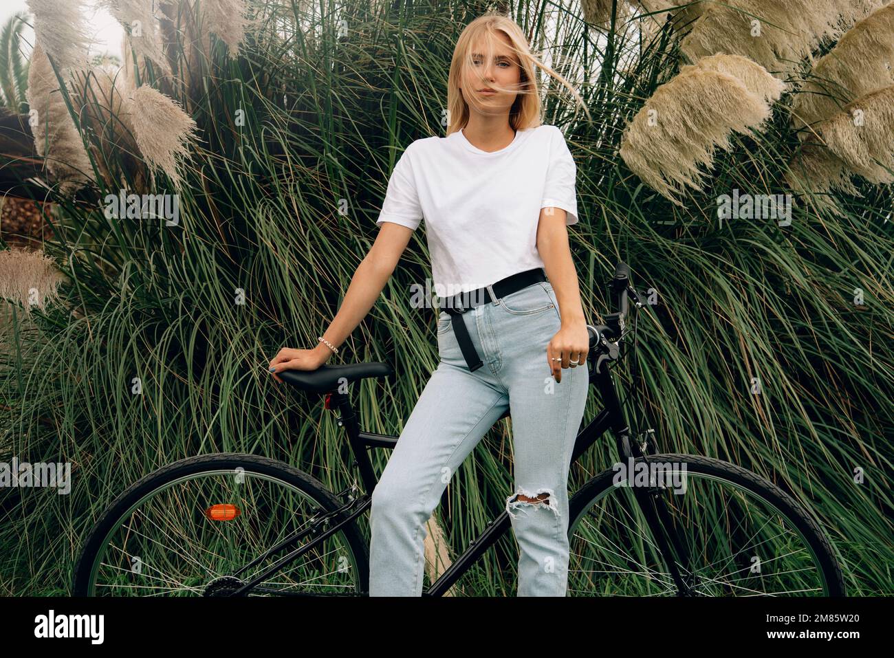 Joven hermosa chica está posando en una camiseta blanca al lado de su bicicleta. Maqueta horizontal. Foto de stock