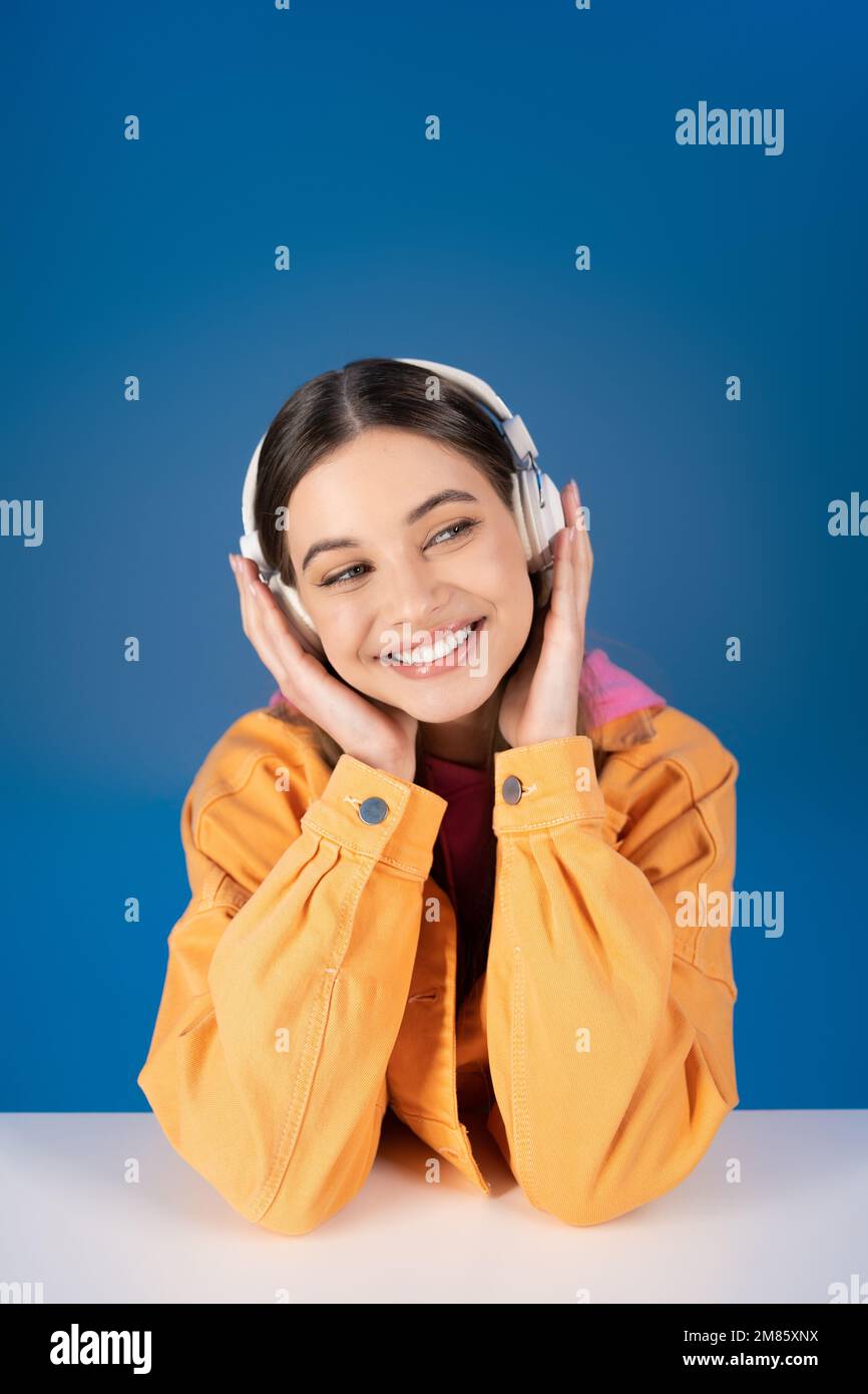 Adolescente positivo en auriculares mirando lejos cerca de la mesa aislada en azul Foto de stock