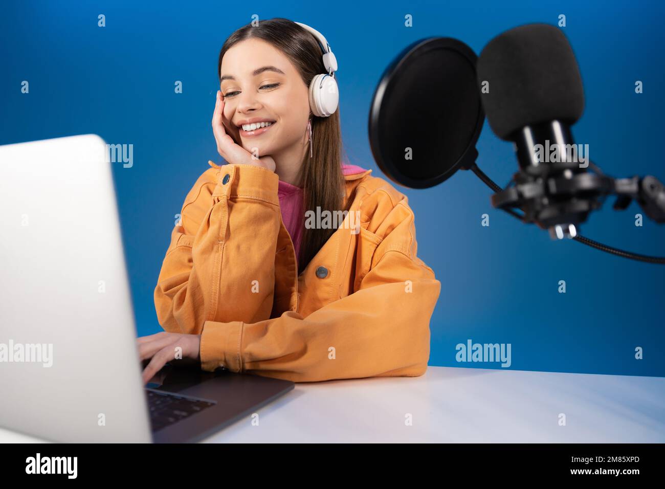 Adolescente en auriculares haciendo podcast cerca de la computadora portátil y el micrófono aislados en azul Foto de stock