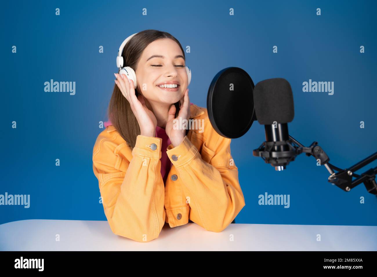 Adolescente complacido en auriculares sentado cerca del micrófono de estudio aislado en azul Foto de stock