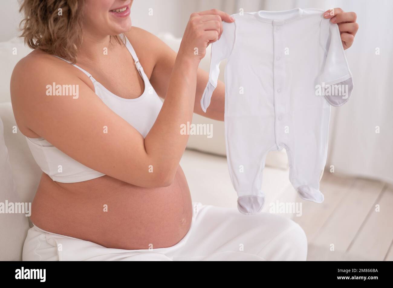 Una mujer embarazada se sienta en un sofá blanco y sostiene la ropa para el bebé esperado. Foto de stock