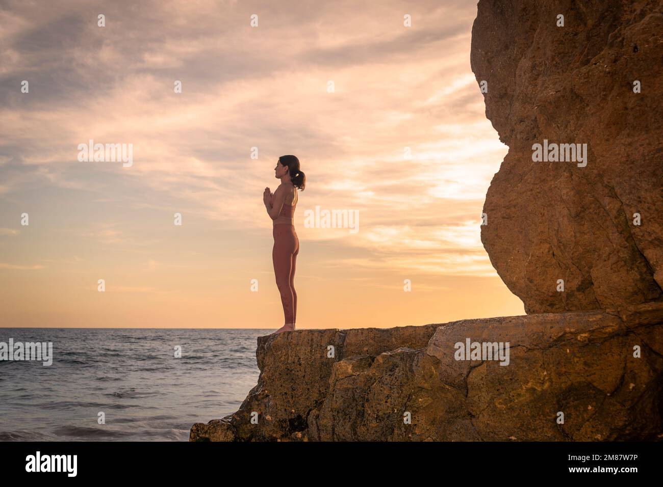 Mujer de pie en las rocas junto al mar practicando yoga al amanecer o al atardecer. Foto de stock