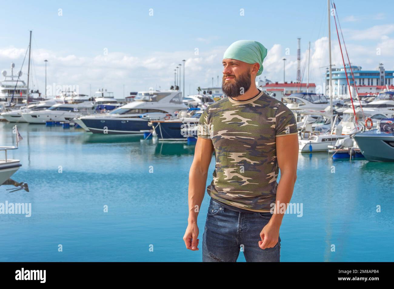 Hermosas camisetas militares bandanna turquesa barba masculina se coloca en el muelle contra el telón de fondo de yates en la bahía del puerto en clima soleado Foto de stock