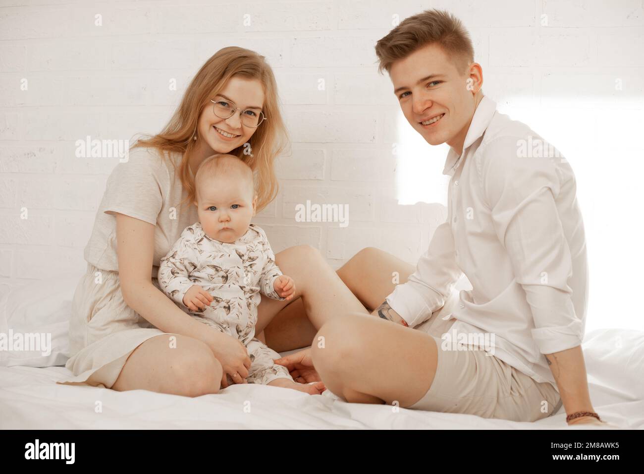 Retrato de familia feliz de tres sobre fondo blanco. Madre, padre y bebé bebé se sientan en la cama y abrazan. Foto de pareja joven y niño en blanco Foto de stock