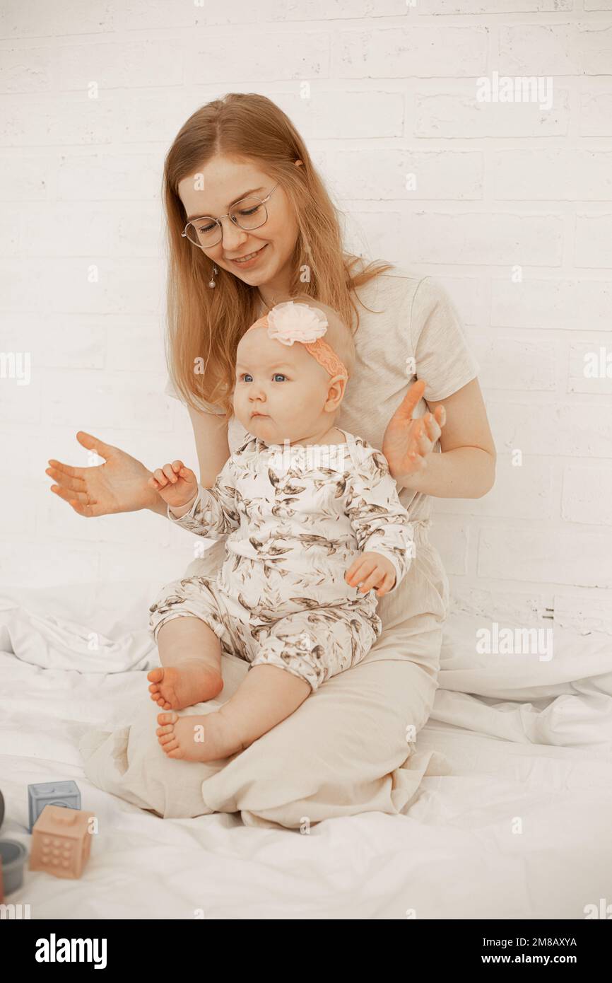 Madre joven feliz con el pequeño bebé se sienta en la cama y juega juguetes sobre fondo blanco. Foto de la familia de la casa de la mujer alegre y del niño lindo del bebé. Concepto Foto de stock