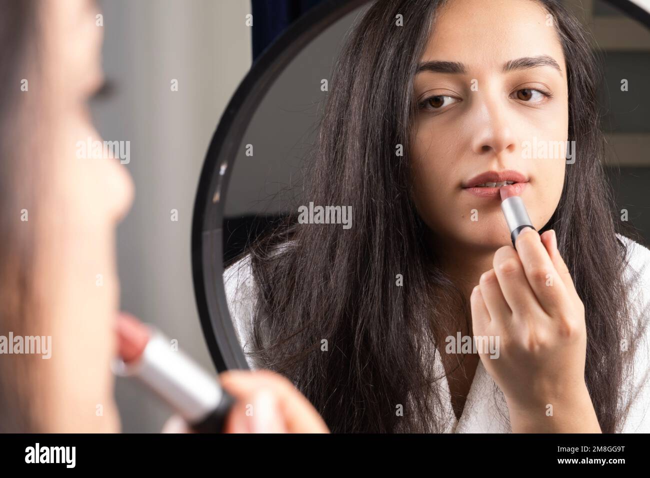 Hermosa mujer milenaria mirando espejo y haciendo maquillaje. Morena chica caucásica rouging lápiz labial. Todos los días rutina de la mañana. Aplicación de cosméticos. Foto de stock