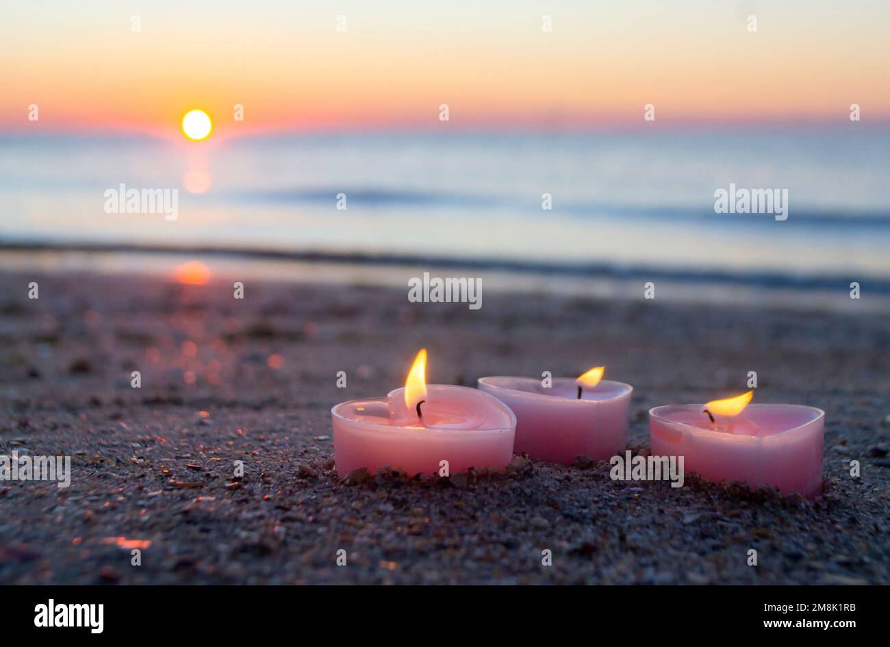 Tres velas en forma de corazón arden en la arena en la playa del mar cerca de las olas del mar contra el fondo del sol brillante durante el amanecer de la puesta del sol. Humor romántico romántico. St Día de San Valentín. Foto de stock