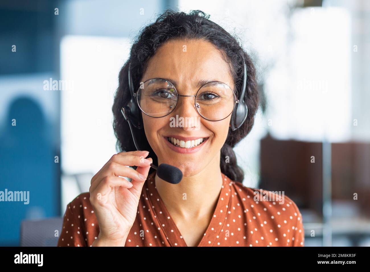 Cierre el retrato de la mujer latinoamericana dentro de la oficina moderna con los auriculares para la videollamada, mujer sonriendo y mirando a la cámara, línea de ayuda de la tecnología de la atención al cliente del trabajador de oficina. Foto de stock