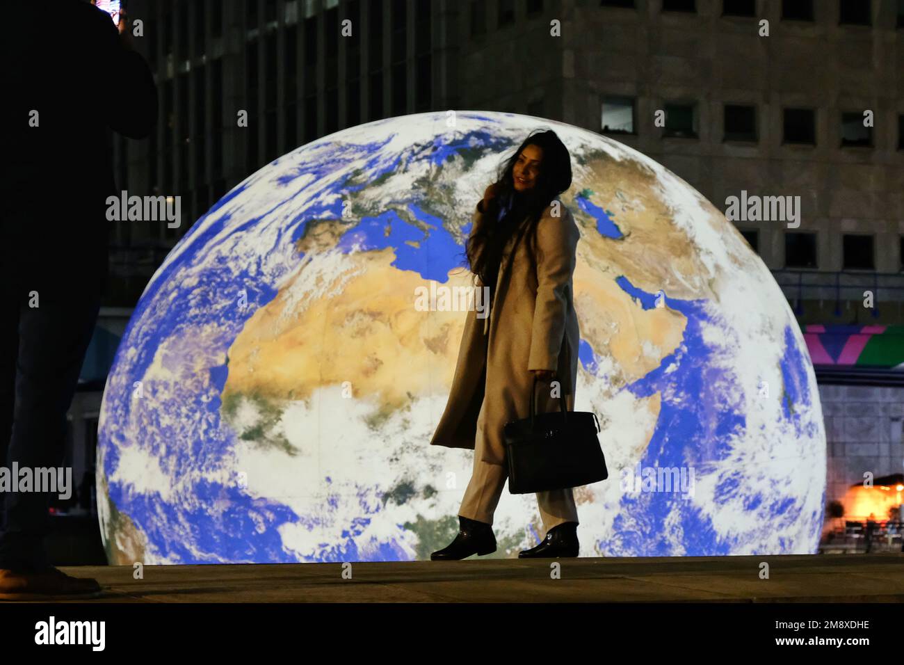 Londres, Reino Unido. 15th de enero de 2023. Una mujer tiene una foto tomada contra un dramático telón de fondo. Floating Earth del artista Luke Jerram se instala antes del Festival de Luces de Invierno de Canary Wharf, que se inaugurará el 18th de enero. La réplica de la tierra iluminada internamente que abarca diez metros de diámetro ha sido creada a partir de imágenes de la NASA vistas desde el espacio, e invita al espectador a reconocer la fragilidad del planeta y considerar las preocupaciones ambientales. Crédito: Undécima hora de fotografía/Alamy Live News Foto de stock