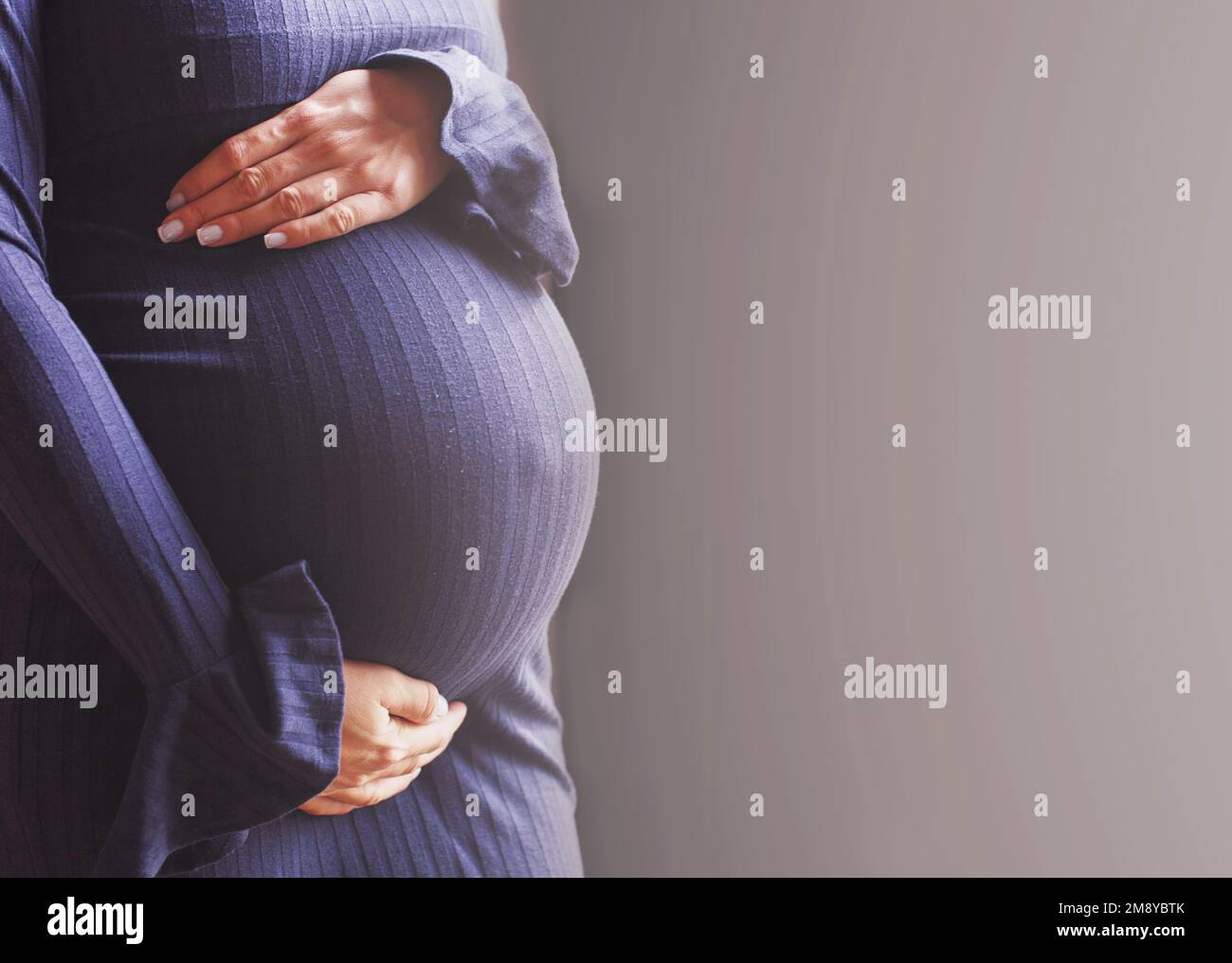 cierre del vientre de una mujer embarazada. Manos de las muchachas abrazando su vientre en fondo azul. Concepto de salud materna. Foto de stock