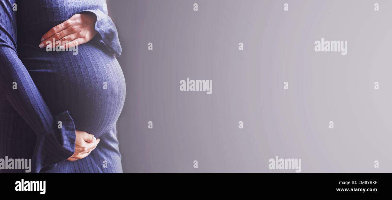 cierre del vientre de una mujer embarazada. Manos de las muchachas abrazando su vientre en fondo azul. Concepto de salud materna. banner Foto de stock