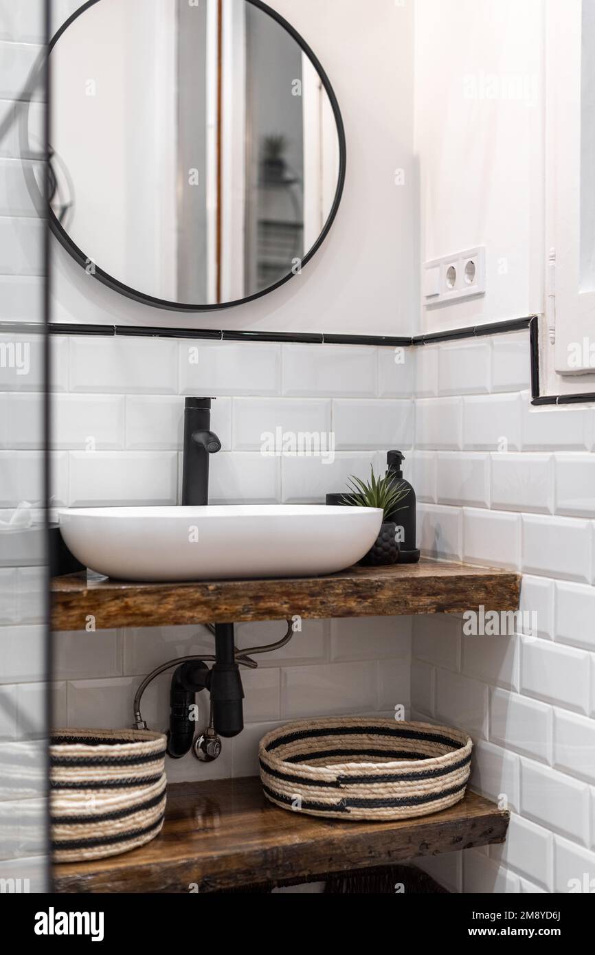 Cuarto de baño con paredes de ladrillo blanco, un lavabo ovalado en una  parte superior de mármol de estilo madera, un espejo redondo en una pared  de marco oscuro refleja una puerta.