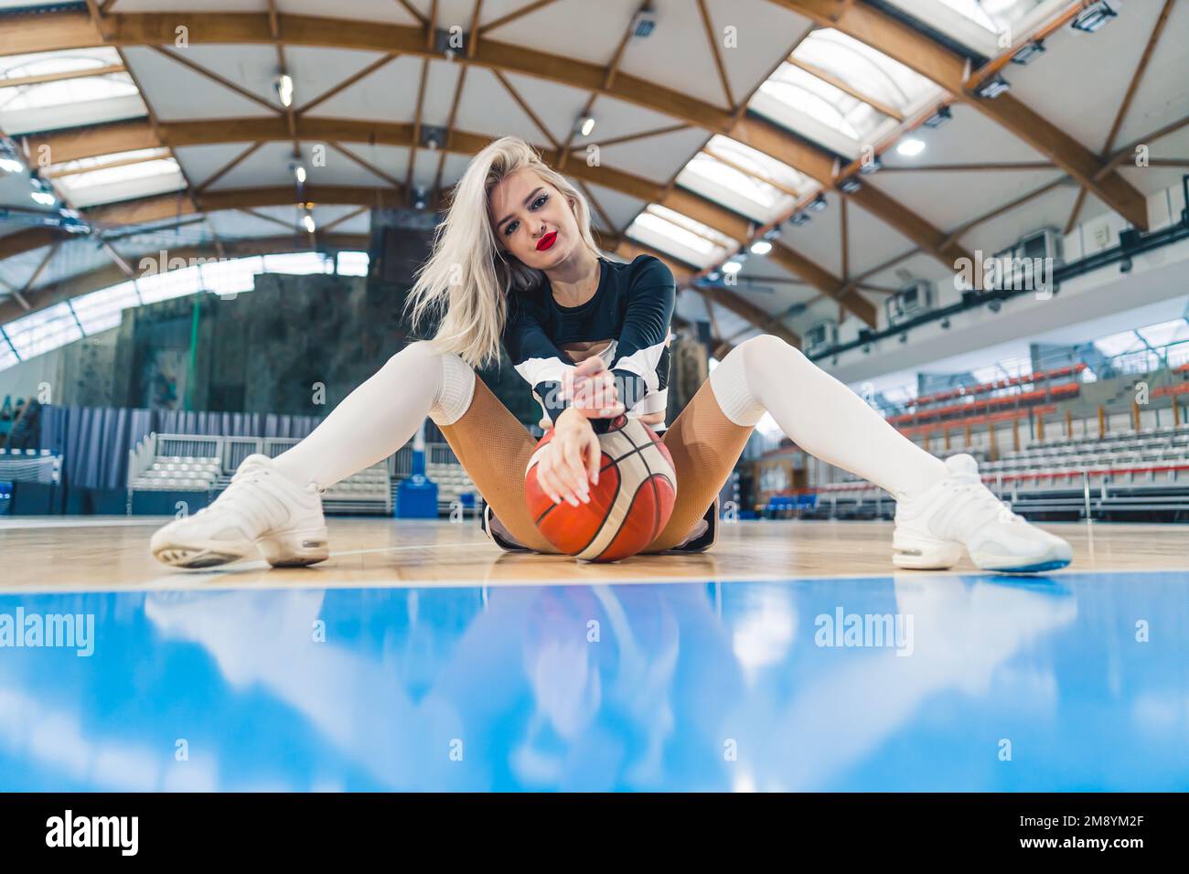 Tiro de ángulo bajo de una animadora rubia sentada en el suelo con un baloncesto entre sus piernas. Concepto deportivo. Foto de alta calidad Foto de stock