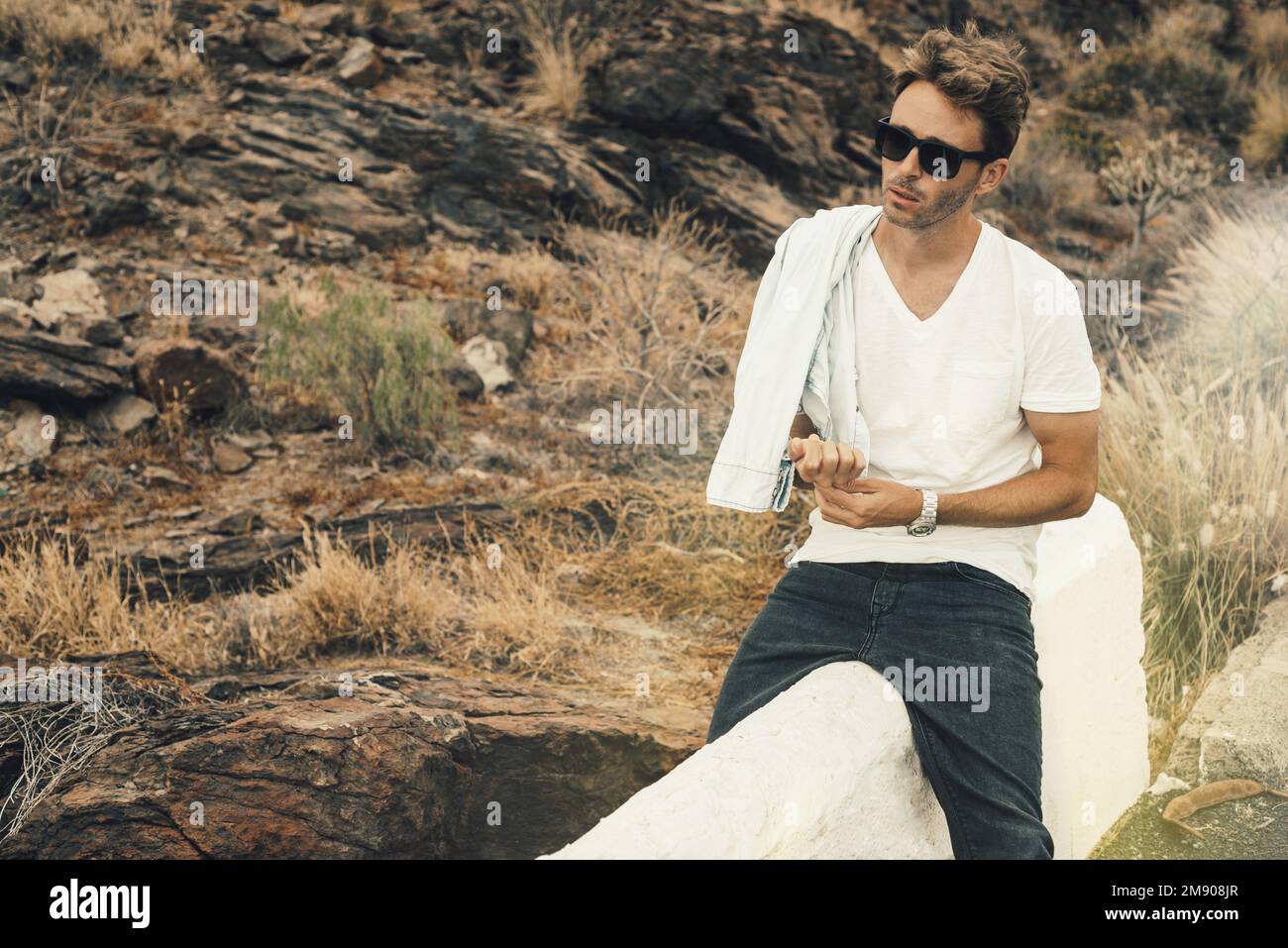 Joven chico guapo sentado en parapeto por la colina rocosa con gafas, camiseta blanca en blanco y jeans negros. Foto de stock