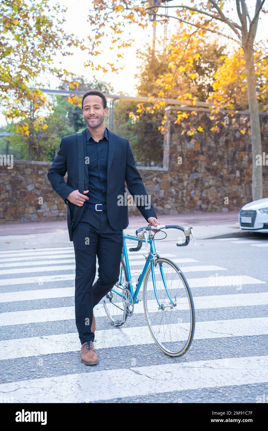 Elegante hombre de negocios afroamericano en traje cruzando la calle con la bicicleta. Foto de stock