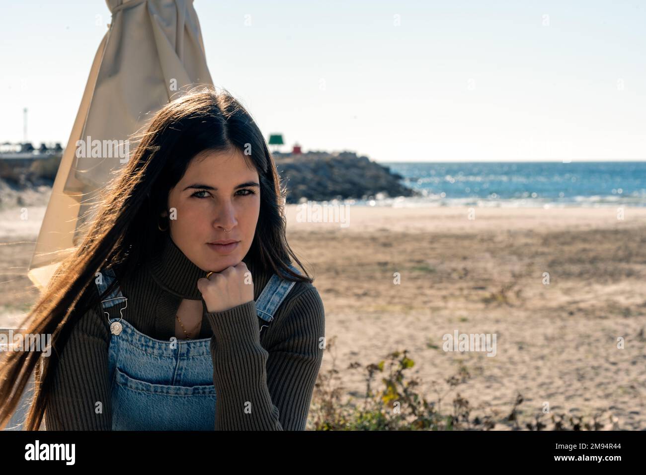 Mujer joven tranquila en denim en general sentado en la playa de arena con la mano en la barbilla contra el mar mirando a la cámara Foto de stock