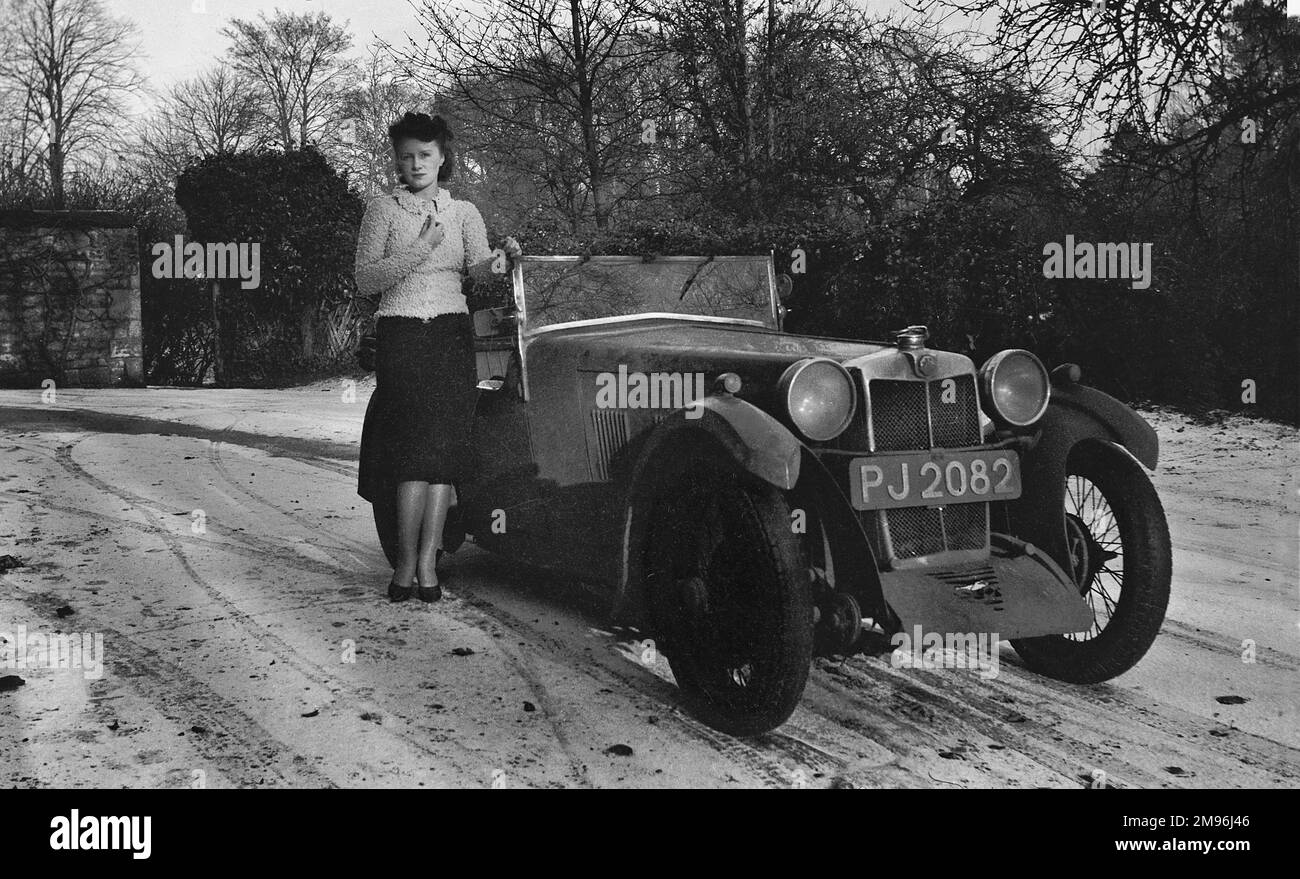 Una mujer joven de pie junto a un coche de techo abierto en clima frío con una salpicadura de nieve en el suelo. Foto de stock