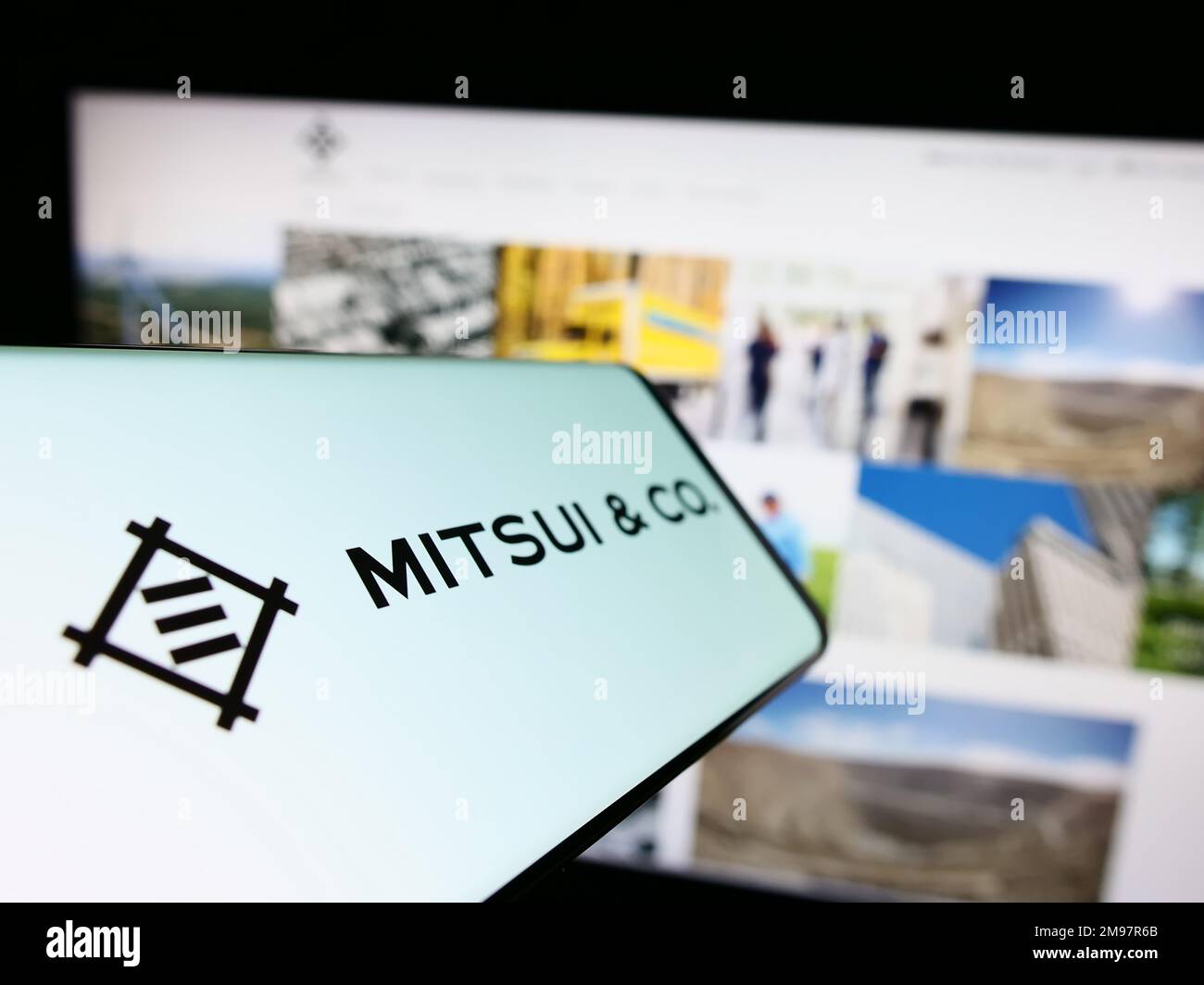 Teléfono móvil con logotipo del conglomerado japonés Mitsui Co. Ltd. En la pantalla frente al sitio web de la empresa. Enfoque en el centro izquierdo de la pantalla del teléfono. Foto de stock