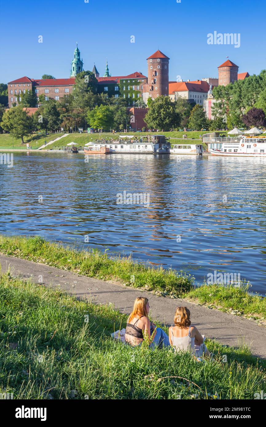 Mujeres jóvenes sentadas en la hierba y disfrutando de la vista sobre el castillo de Wawel en el río en Cracovia, Polonia Foto de stock