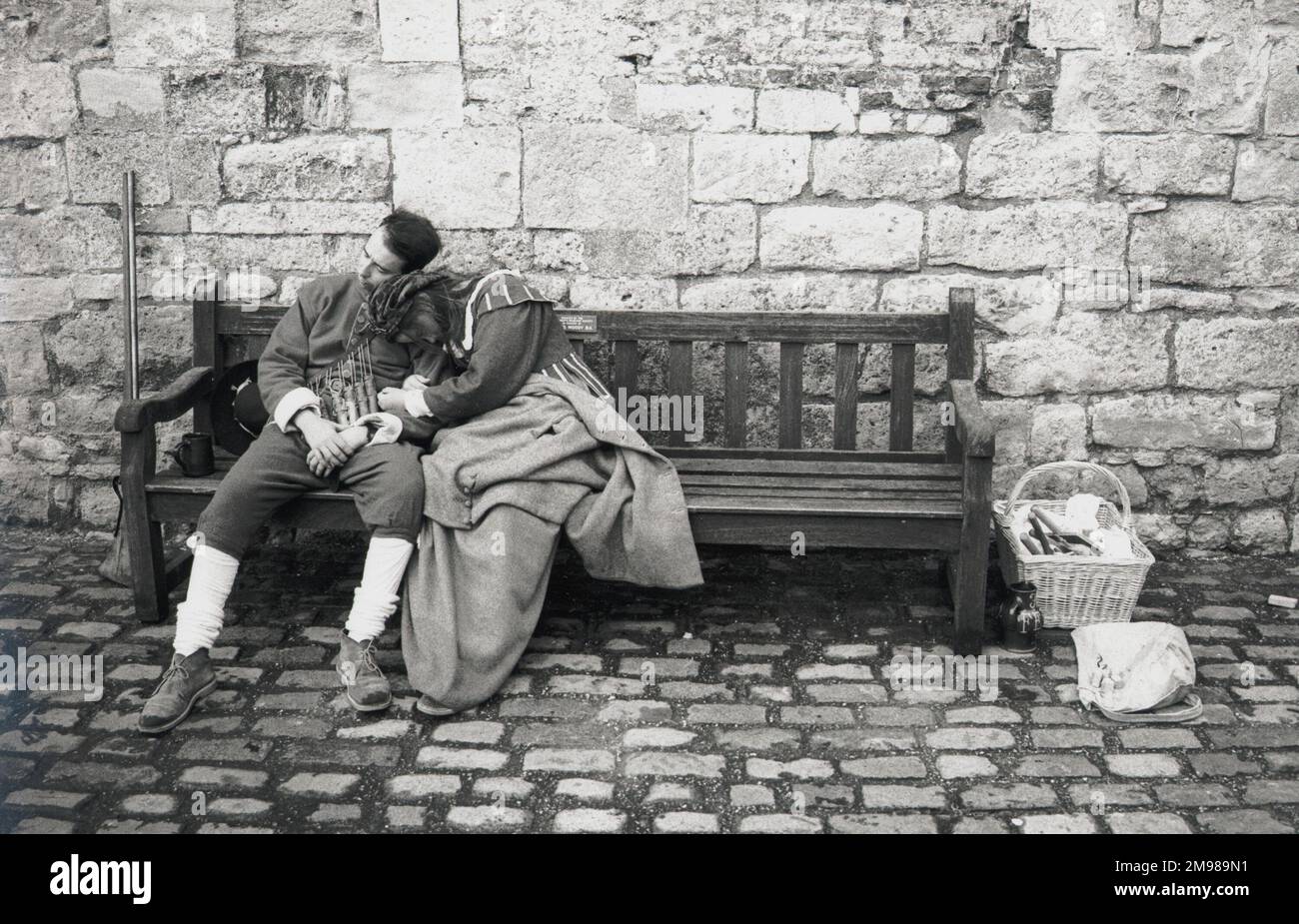 Una pareja vestida con traje de época, durmiendo en un banco en un patio empedrado. Foto de stock