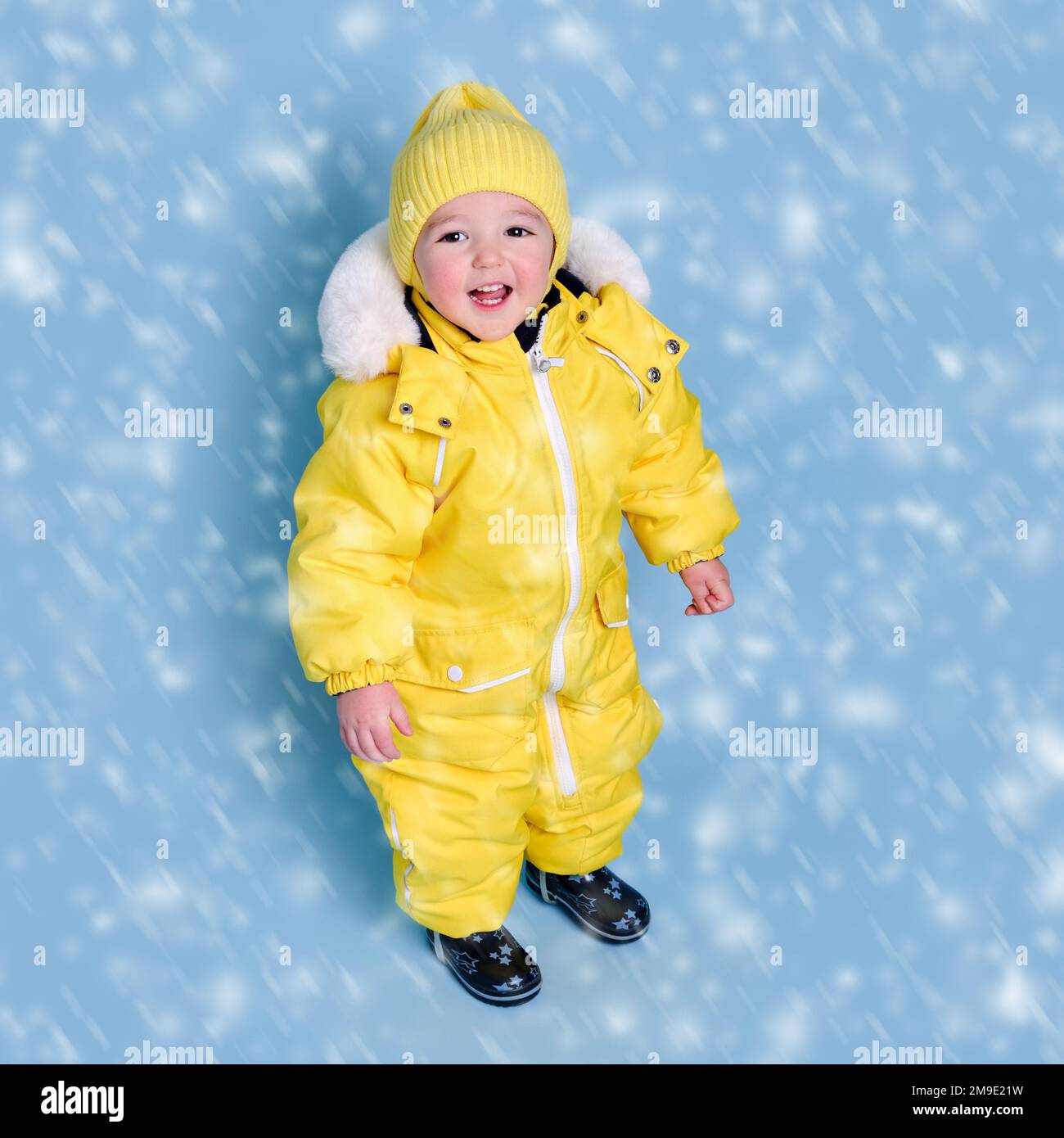 Feliz bebé en invierno traje de nieve ropa en estudio fondo azul. Un niño  en un traje amarillo cálido con capucha. Niño de un año y cinco meses  Fotografía de stock -