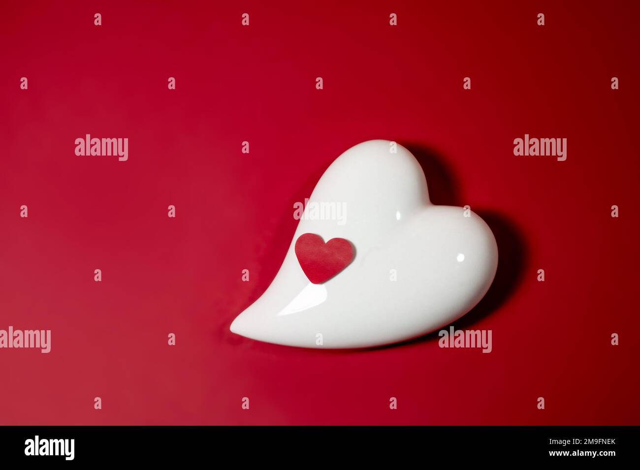 Corazón de porcelana blanca y pequeño corazón de papel sobre un fondo rojo, símbolo de amor y tarjeta de felicitación para vacaciones como San Valentín o el día de las madres, espacio de copia Foto de stock