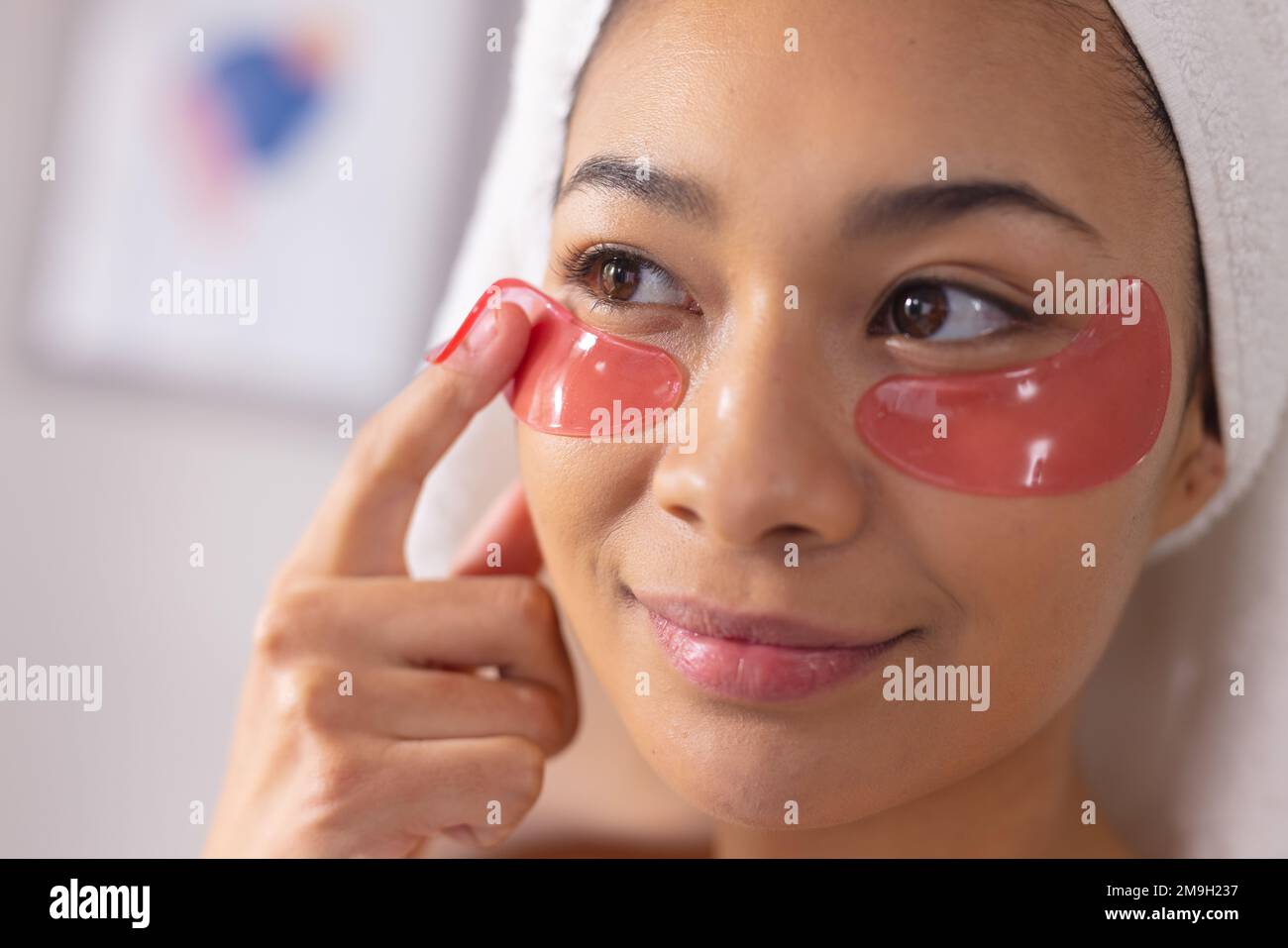 Imagen de mujer biracial con toalla en la cabeza aplicando máscara de ojos en el baño Foto de stock