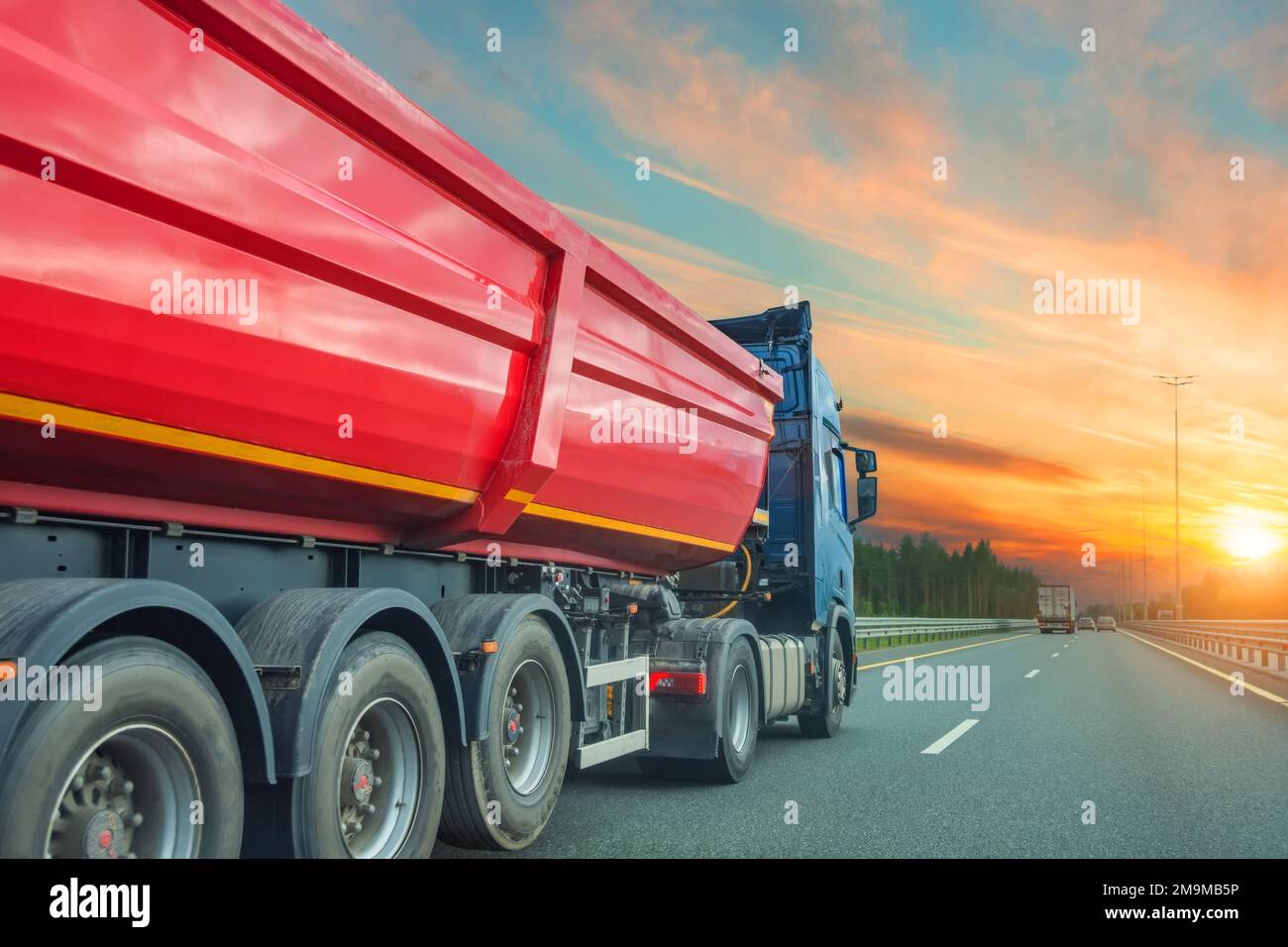 El remolque rojo grande del camión de volteo monta en la carretera, en el cielo la puesta del sol brilla sobre el horizonte Foto de stock