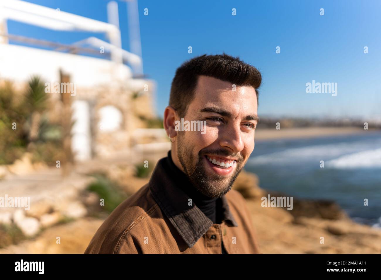 Macho barbudo hispano positivo mirando hacia otro lado en el fondo borroso del edificio envejecido situado en la orilla del mar en el sol Foto de stock