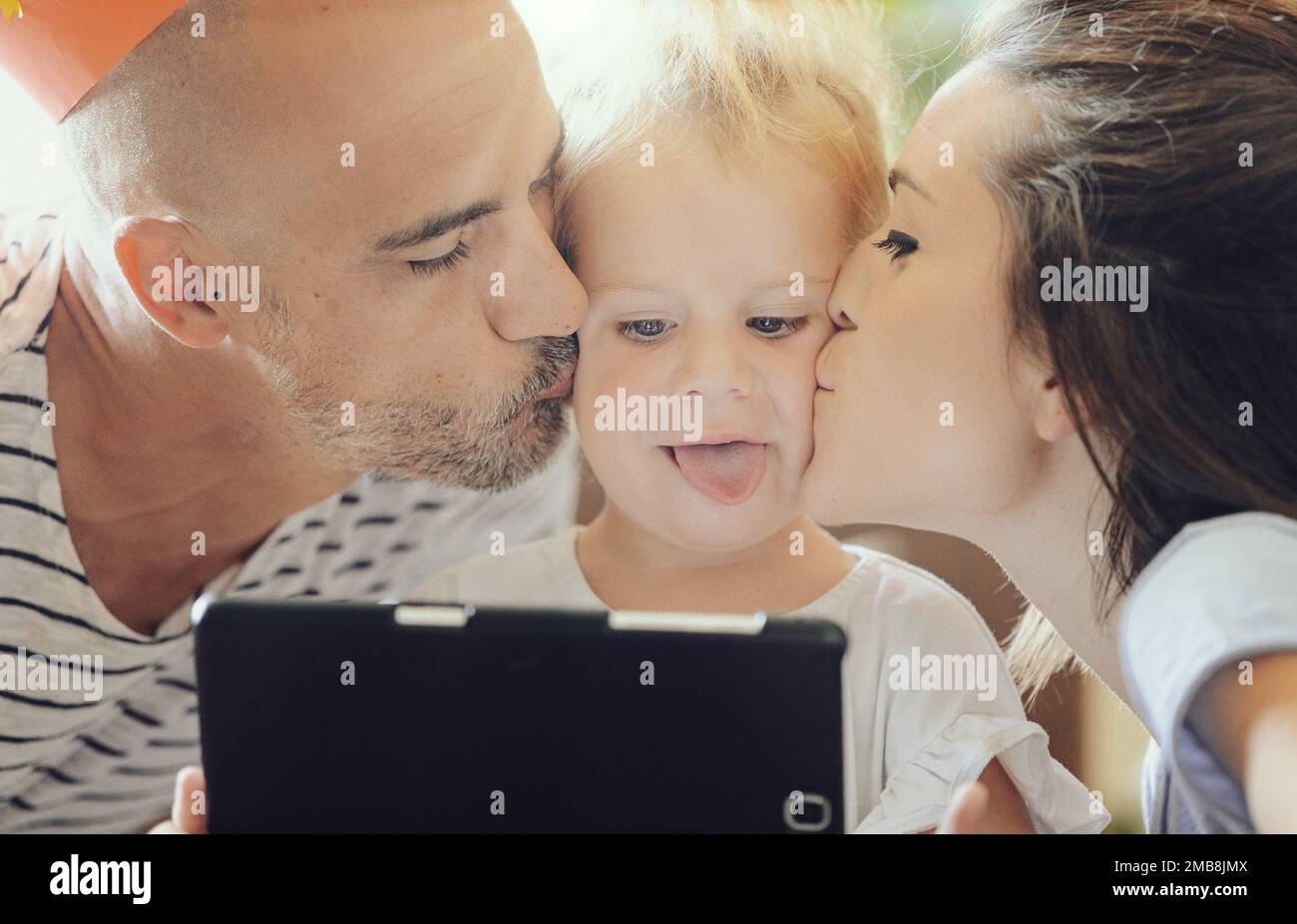 Foto de primer plano de dos padres besando a su niña en sus mejillas mientras juega con la tableta (se mete la lengua) Foto de stock