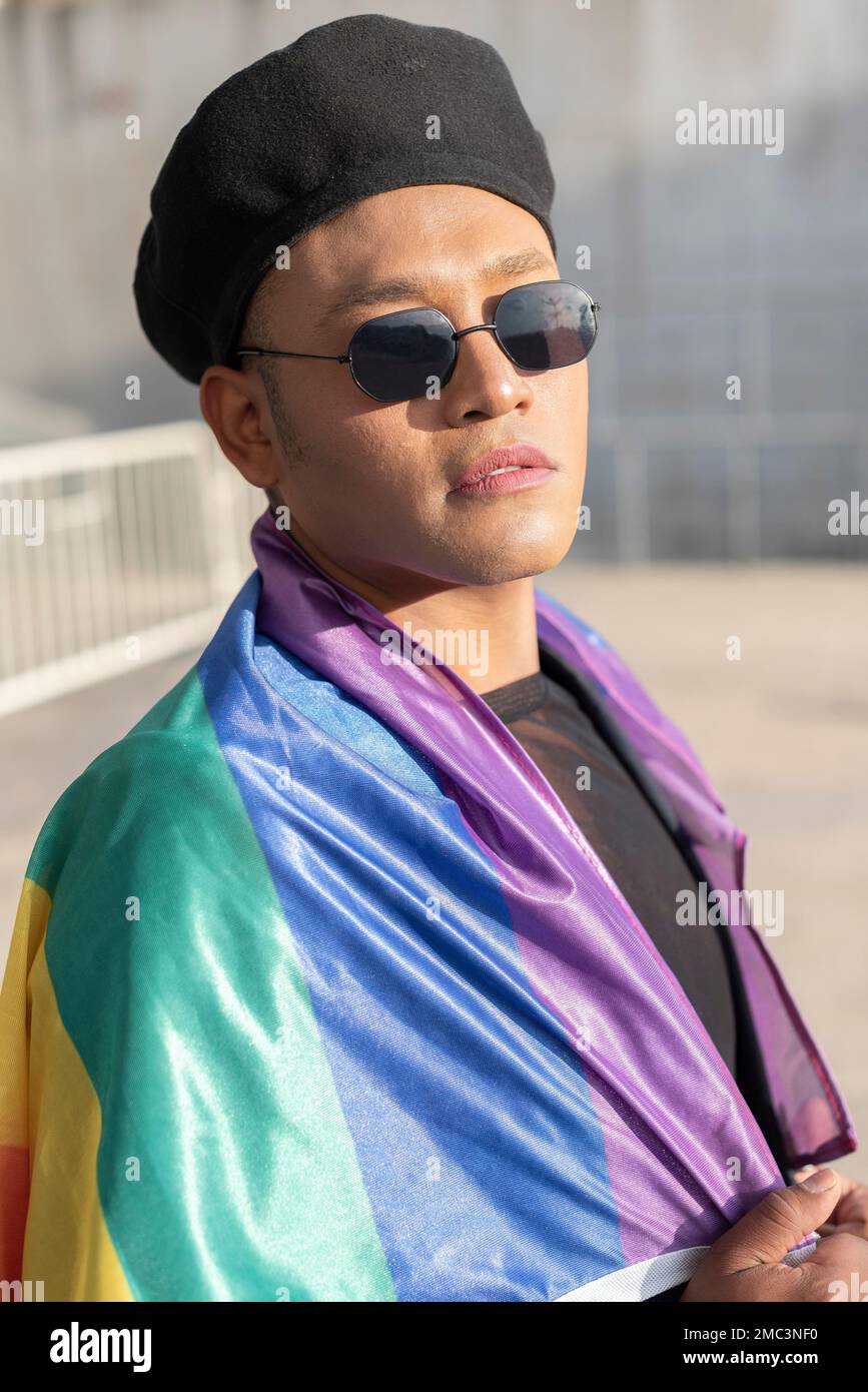 Hombre gay con maquillaje en debajo de la bandera del arco iris LGBT mientras mira a la cámara. Concepto de orgullo. Mes del orgullo. Foto de stock