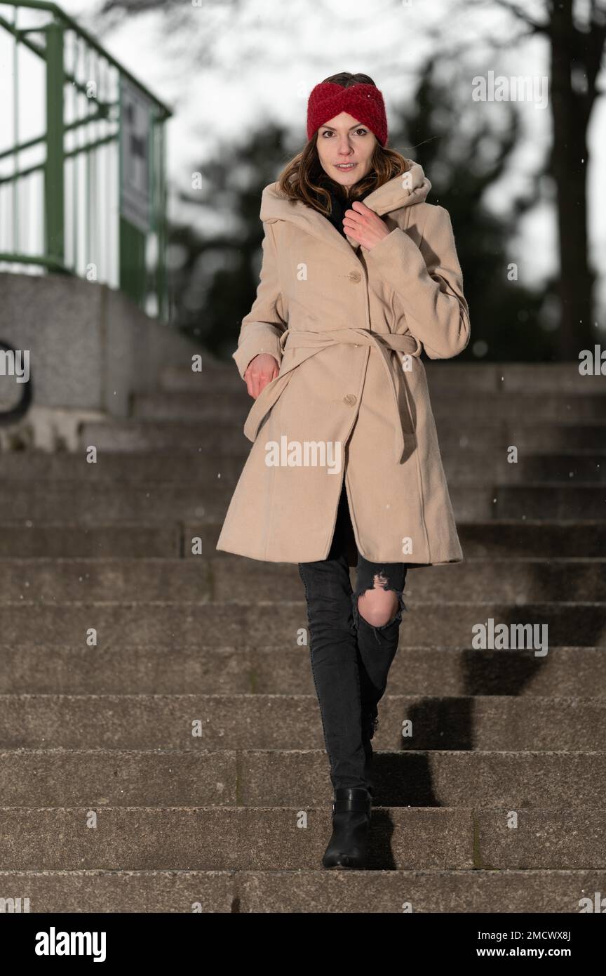 Mujer joven bonita sonriente con el pelo largo moreno, un abrigo de tela  marrón claro y un gorro de lana roja camina por una escalera de piedras  naturales Fotografía de stock -