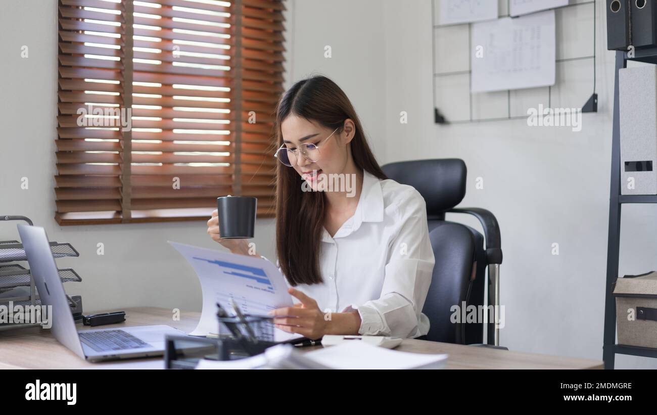 Concepto de negocio, empresaria está leyendo el documento financiero y tomando café en la oficina moderna. Foto de stock