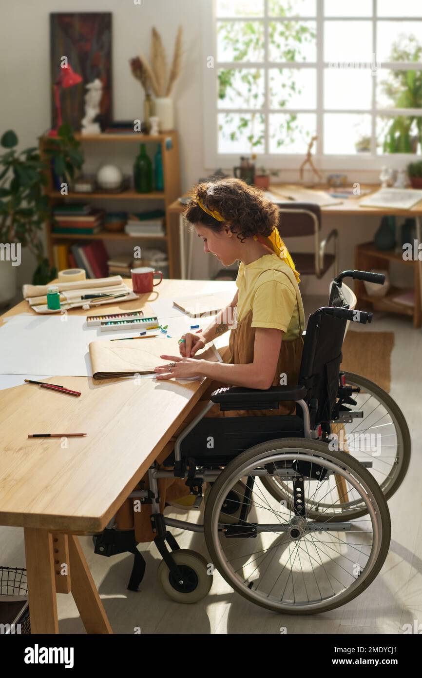 Muchacha adolescente en silla de ruedas sentada junto a la mesa de madera con suministros para la creación de arte en su estudio casero y dibujo nueva obra maestra Foto de stock