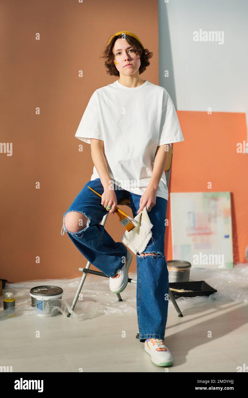 Joven pintora creativa en ropa casual sosteniendo pinceles mientras se sienta en la silla delante de la cámara con el pedazo de cartón detrás Foto de stock